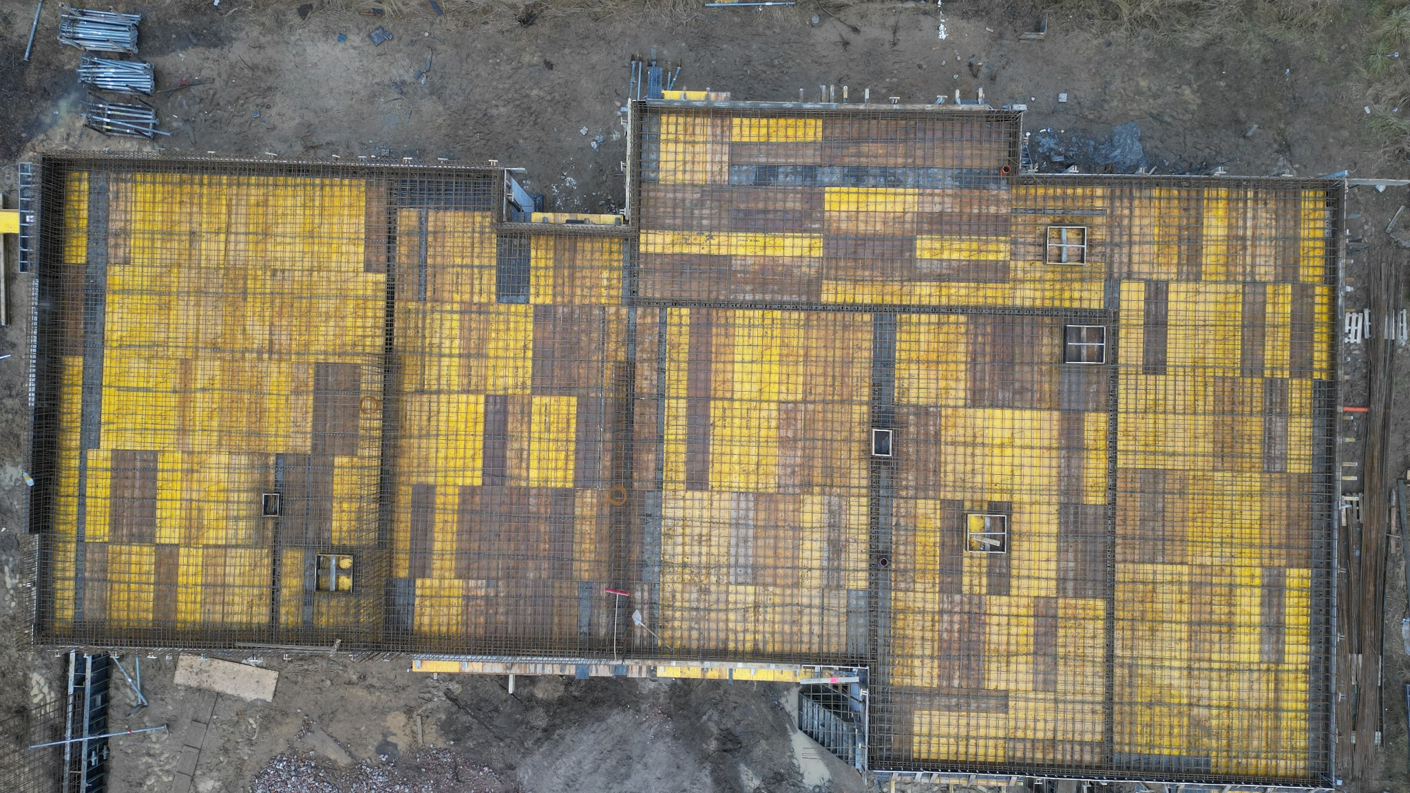 Widok z góry na deskowanie i zbrojenie fundamentów budynku z widocznymi otworami okiennymi w ziemi, przygotowane do wylania betonu.