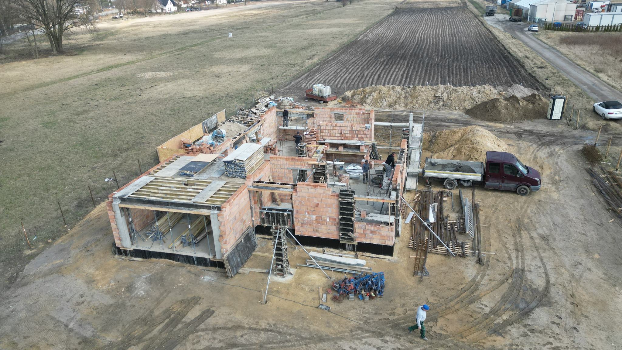 Widok z góry na plac budowy domu jednorodzinnego w stanie surowym otwartym, z widocznymi murami z czerwonej cegły, częściowo zaszalowanymi elementami żelbetowymi, rusztowaniami, pracownikami...