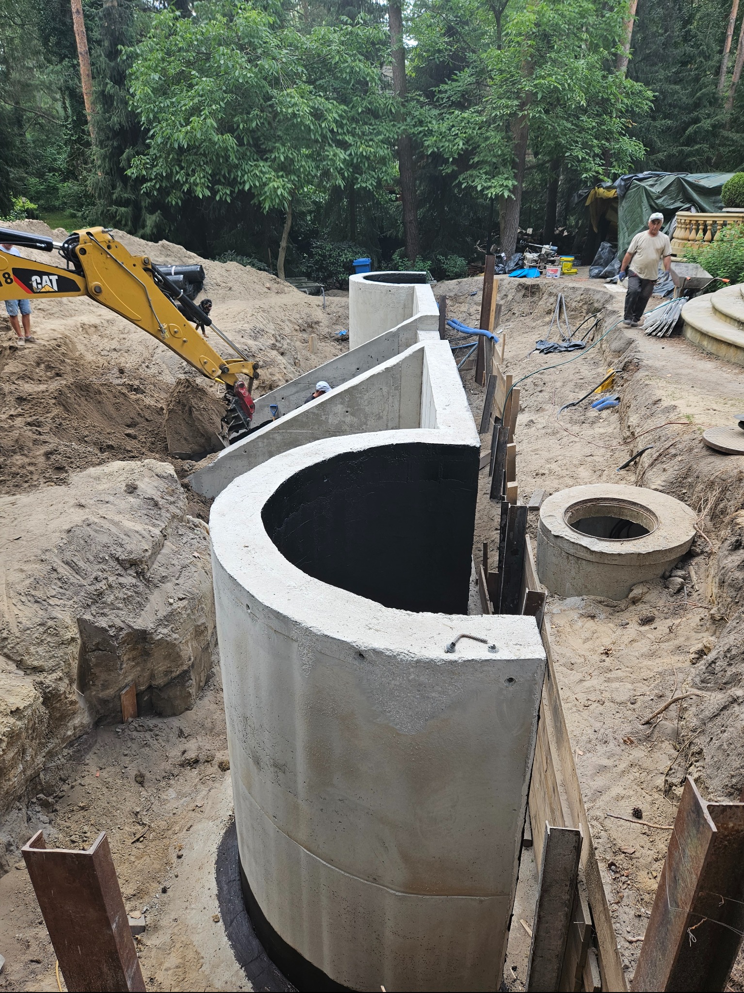 Budowa podziemnego zbiornika retencyjnego z prefabrykatów betonowych, widoczny wykop, zbrojenie, szalunki i pracujący mały koparka w tle, teren zalesiony.