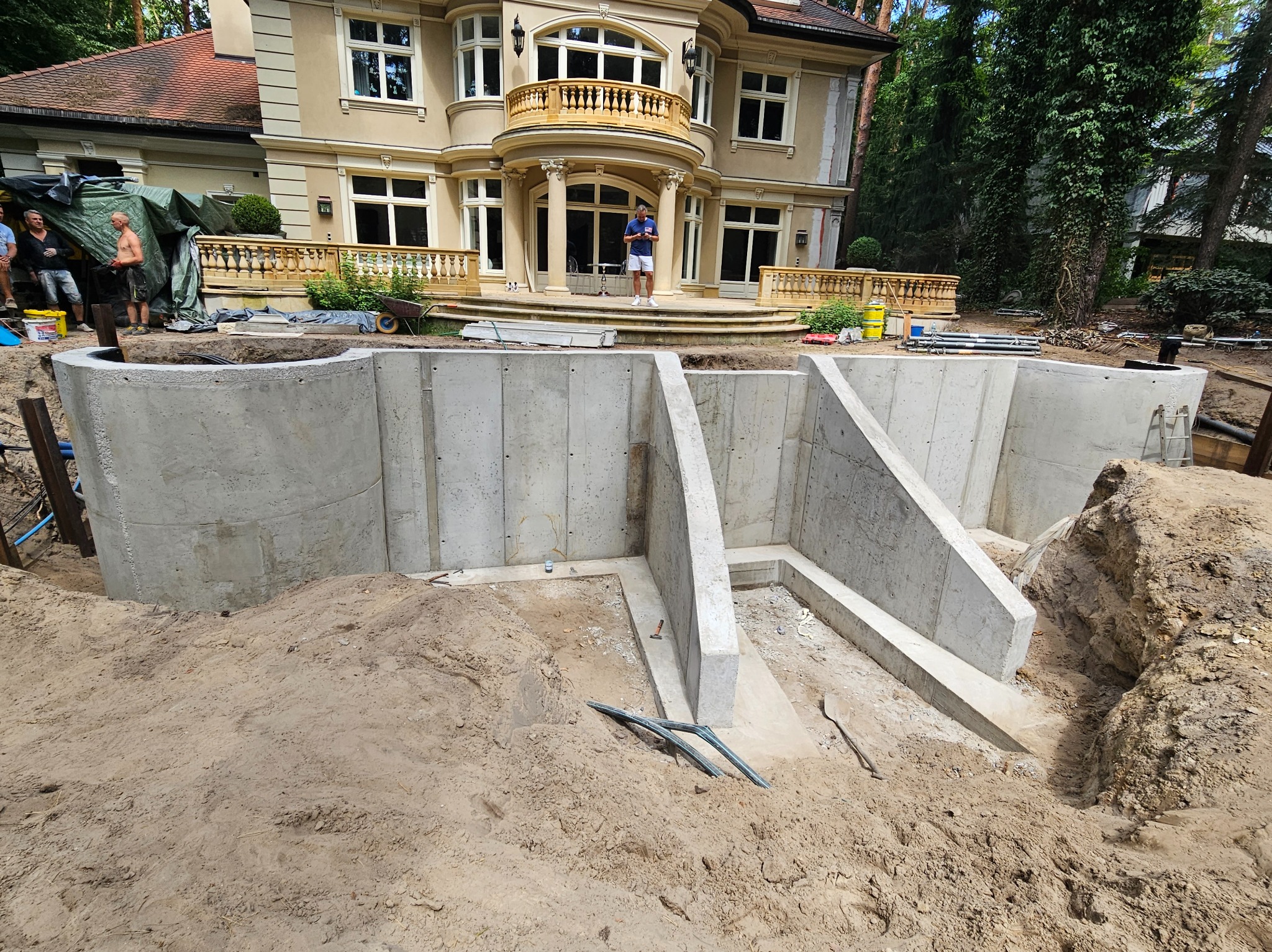 Wykop pod budynek z częściowo wylanymi betonowymi ścianami fundamentowymi, widoczne zbrojenia i deskowania w tle, luksusowa rezydencja w stylu klasycznym z balkonem.