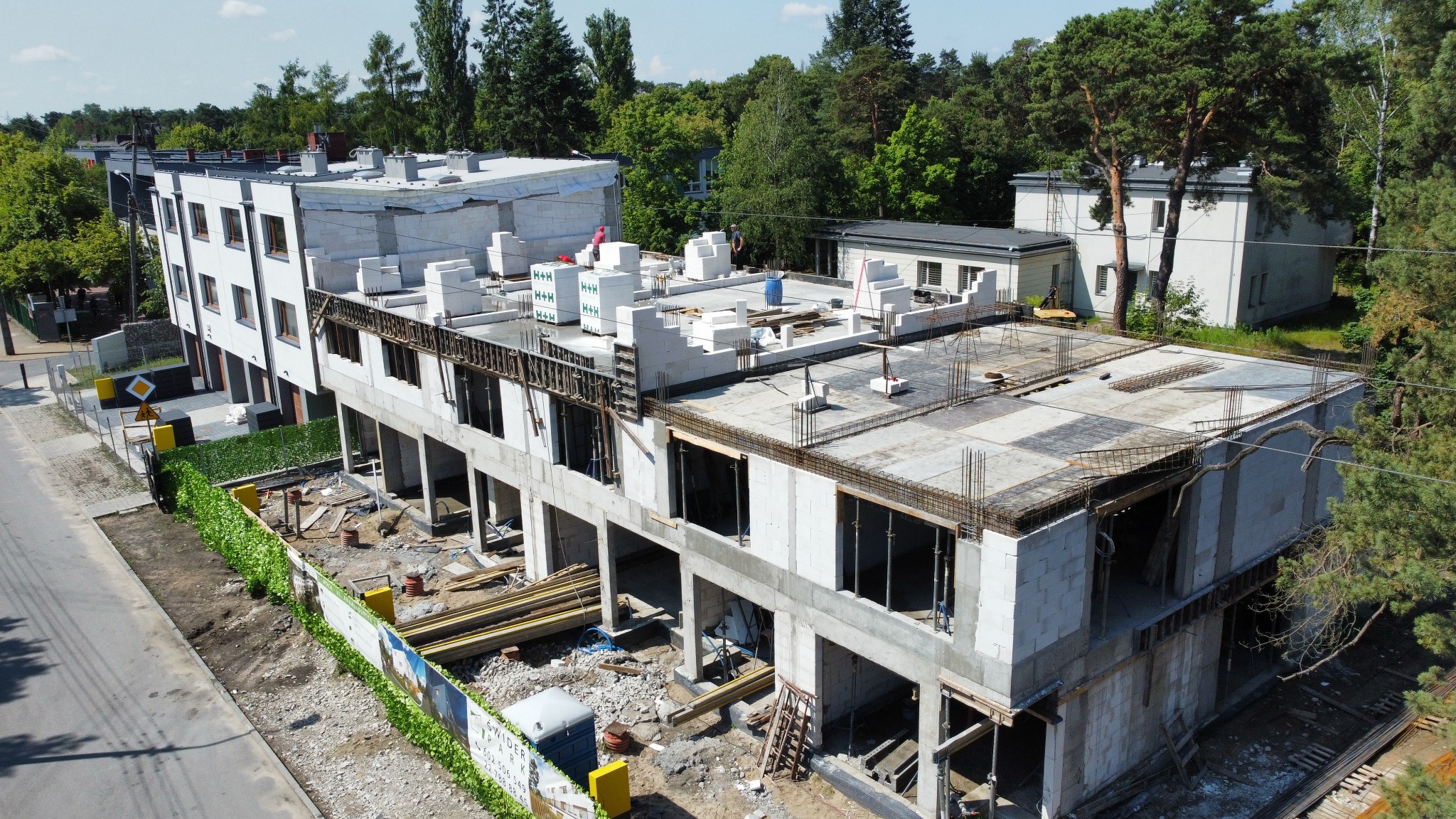 Wysoki kąt widoku na plac budowy z surową konstrukcją wielorodzinnego budynku z betonu komórkowego, widoczne zbrojenia na stropie, palety z bloczkami H+H na dachu i pracownicy w oddali.