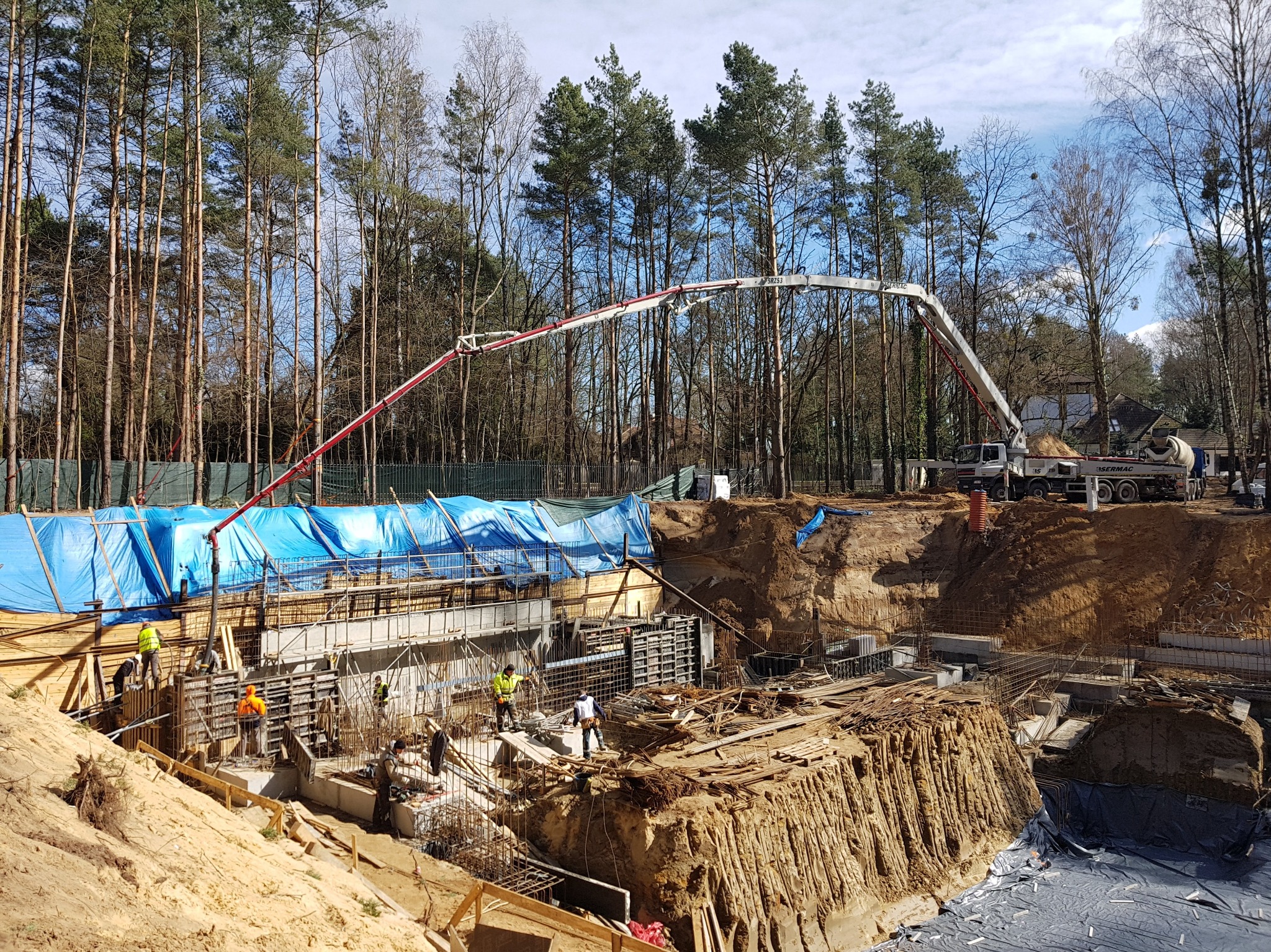 Wykop pod budynek z szalunkami i zbrojeniami, widoczny betonowóz z pompą podającą beton, pracownicy w odblaskowych kamizelkach na placu budowy, otoczenie stanowią drzewa.