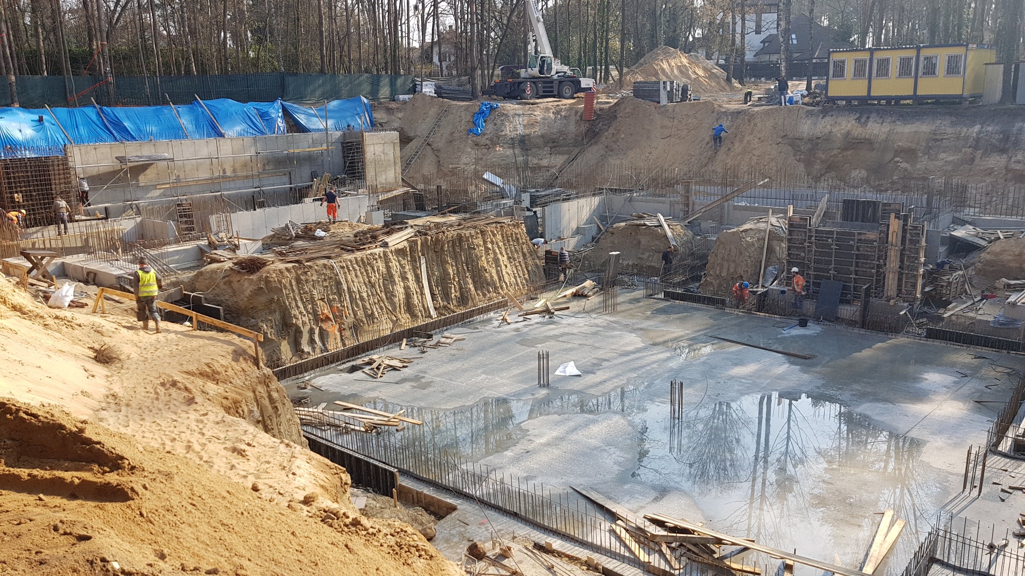 Szeroki kadr na plac budowy z wykopem pod fundamenty, widoczne zbrojenia, szalunki, pracownicy w odblaskowych kamizelkach, żuraw budowlany w tle i częściowo zalany beton