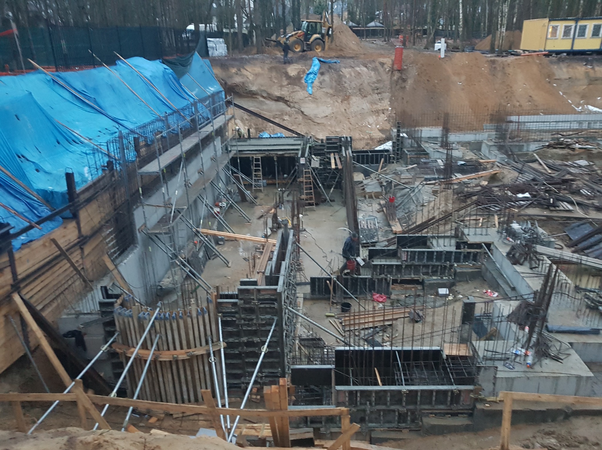 Fundamenty budynku w trakcie budowy: widoczne zbrojenia, szalunki, betonowe elementy oraz pracujący robotnik w kurtce, na tle ziemi, drzew i żółtej barakowozu.