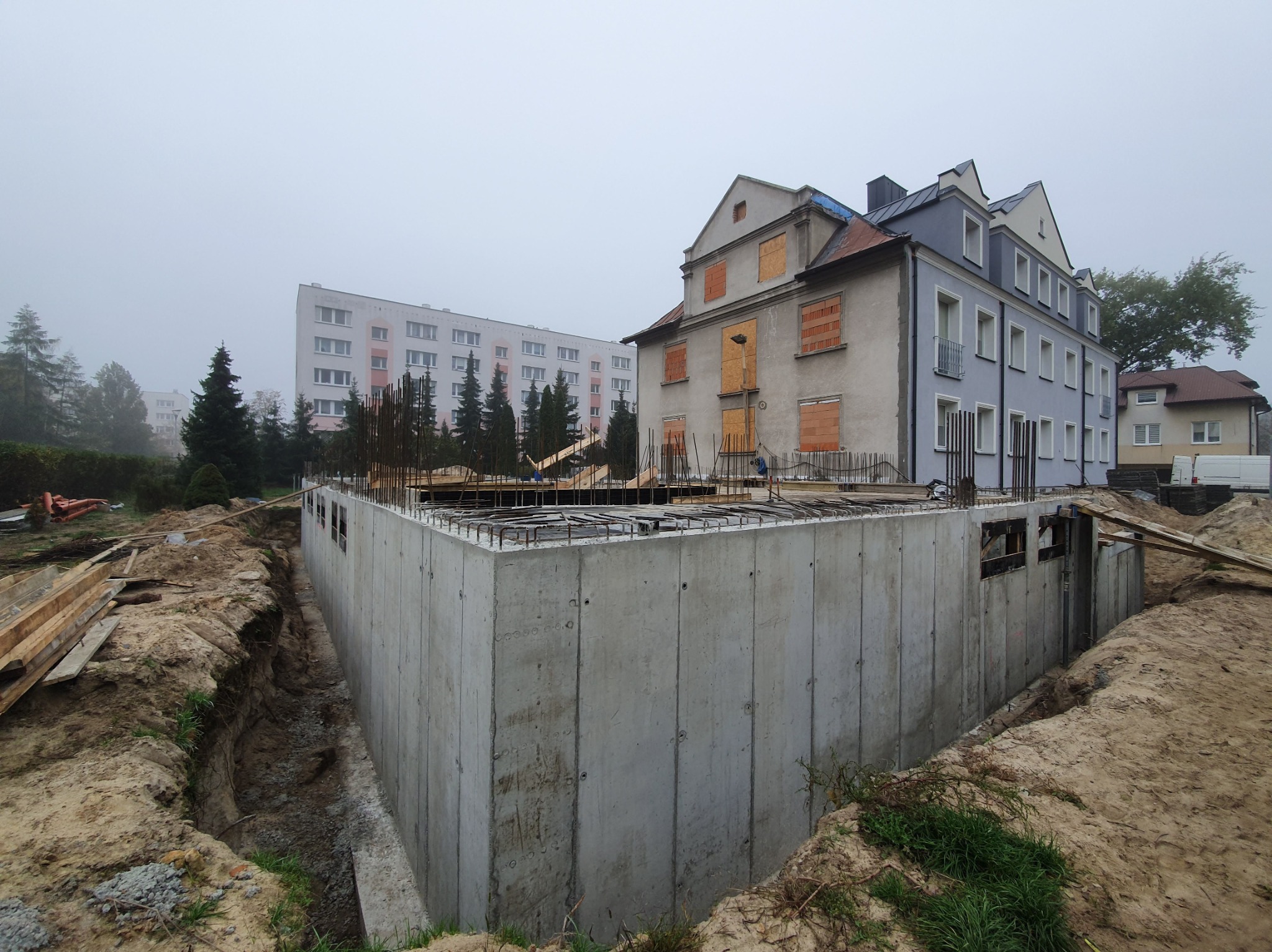 Wylany fundament z betonu zbrojonego pod budynek, widoczne zbrojenie na górze, w tle budynki mieszkalne, pochmurny dzień.