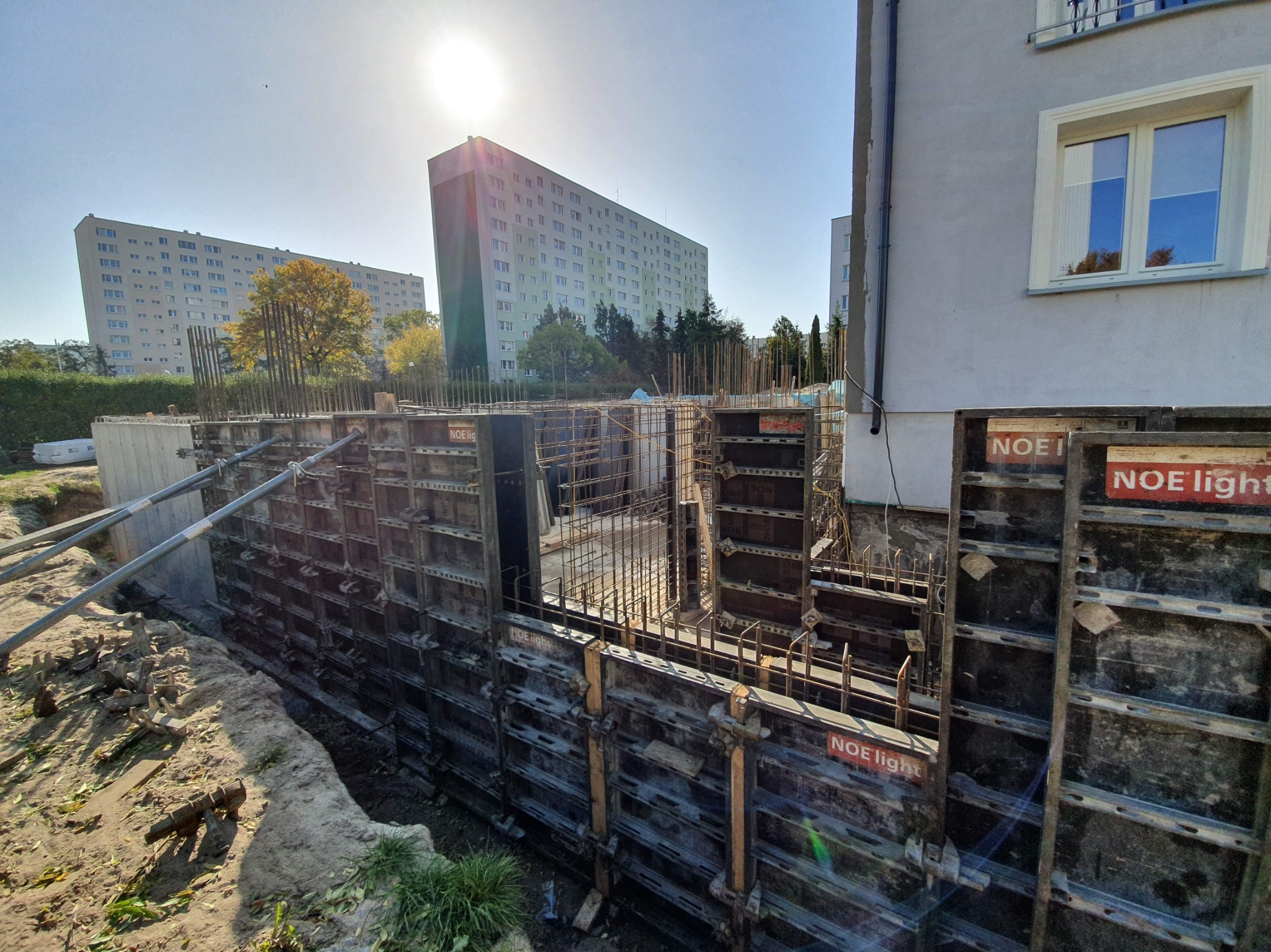 Przygotowanie szalunku i zbrojenia pod mury oporowe przy budynku mieszkalnym, widoczne elementy konstrukcyjne NOE light, w tle bloki mieszkalne i błękitne niebo z promieniami słońca.