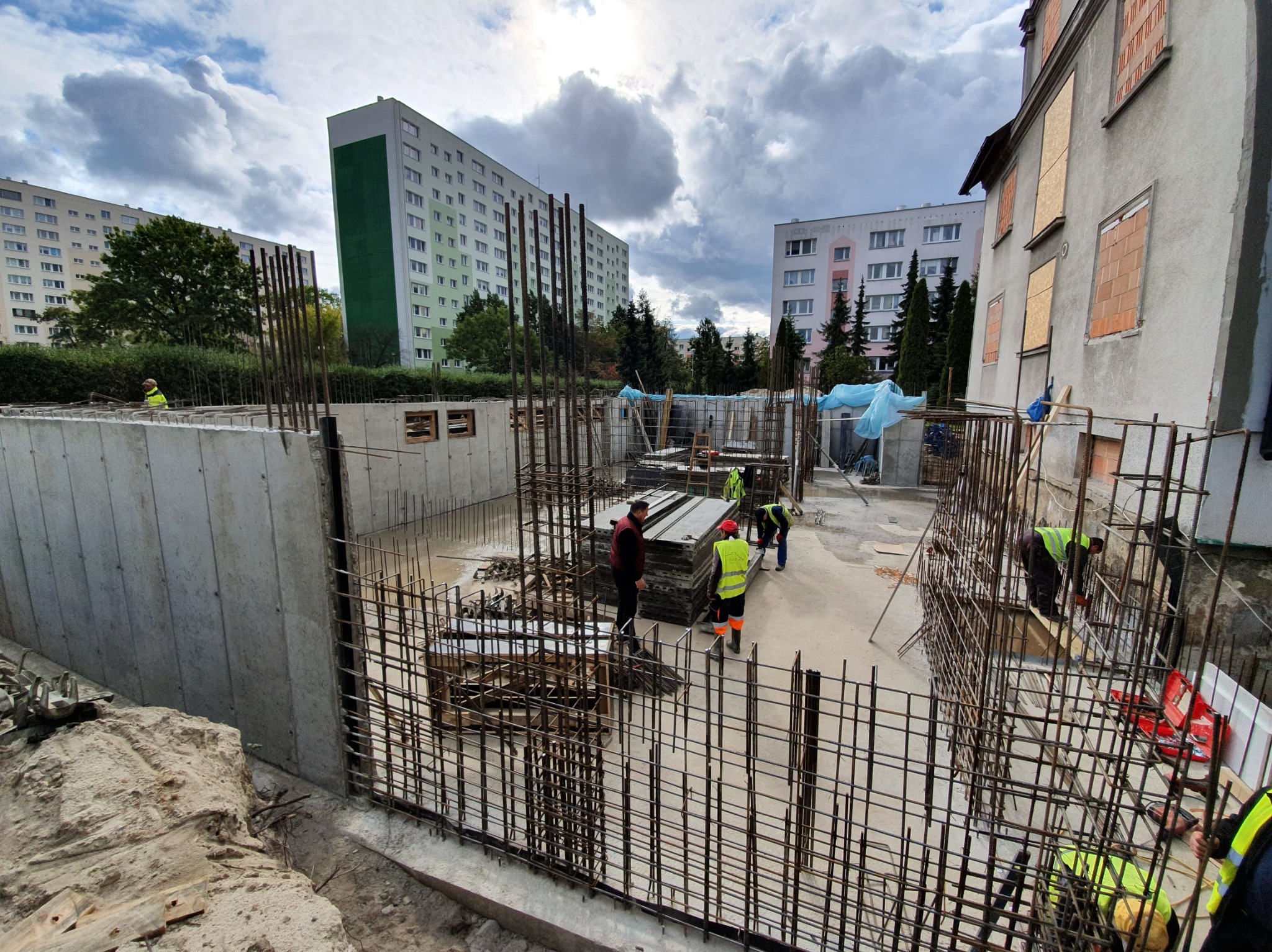 Budowa fundamentów budynku wielorodzinnego: widok na zbrojenie, szalunki i pracujących robotników w odblaskowych kamizelkach na tle bloków mieszkalnych i częściowo zamurowanego budynku.
