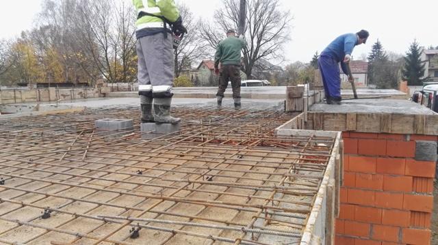 Zbrojenie stropu budynku w trakcie budowy, widoczni robotnicy w roboczych ubraniach i kaloszach, przygotowujący szkielet z prętów zbrojeniowych.