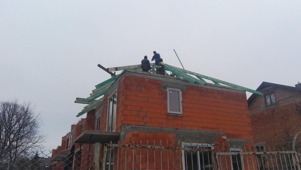 Ekipa budowlana montuje drewnianą konstrukcję dachu na niewykończonym domu z czerwonej cegły, widok z dołu, pochmurne niebo w tle.