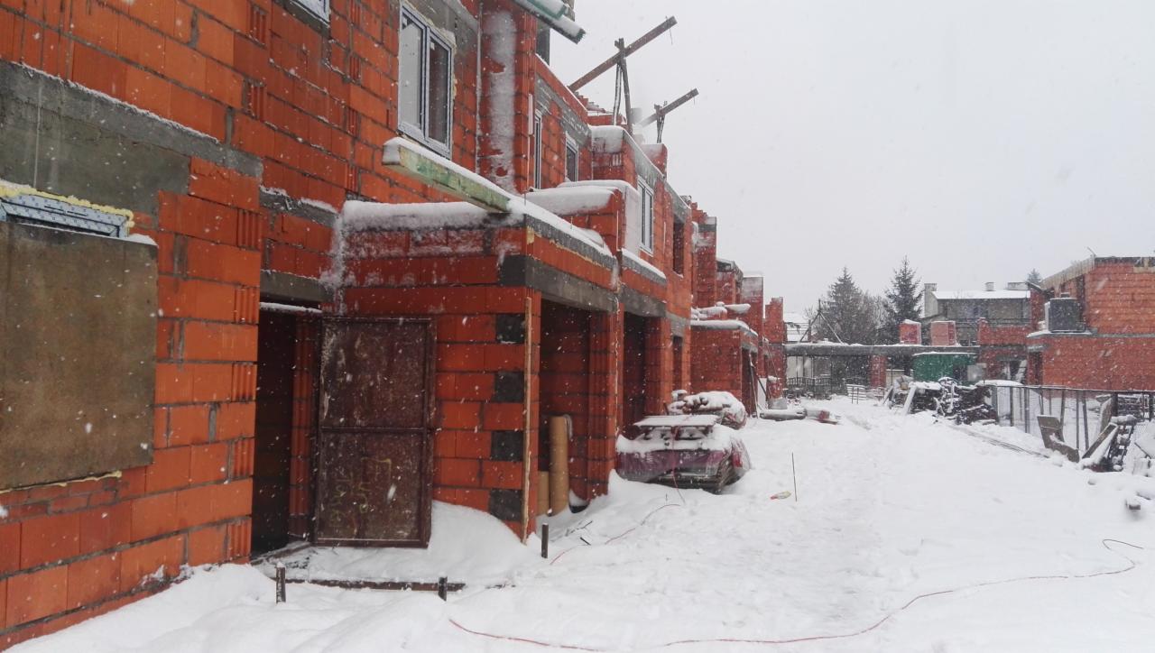 Surowy stan budowy szeregu domów z czerwonej cegły w zimowej scenerii, zasypane śniegiem, widoczne niedokończone garaże i elementy konstrukcyjne.
