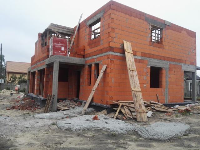 Budowa domu jednorodzinnego w stanie surowym otwartym, widoczne ściany z czerwonej cegły, betonowe słupy i drewniane deski opierające się o ściany, na pierwszym planie gruz i piasek.