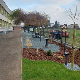 KOLOROWY OGRÓD SPÓŁKA Z OGRANICZONĄ ODPOWIEDZIALNOŚCIĄ - Uporządkowany teren zielony przy budynku szkolnym z elementami dekoracyjnymi: kora, kamień ozdobny, lampy solarne, karmnik dla ptaków, z tyłu widoczny traktor z przyczepą.