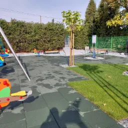 Nowo założony trawnik z rolki graniczący z placem zabaw wyłożonym gumową nawierzchnią, młode drzewko zabezpieczone palikami, w tle żywopłot i elementy placu zabaw.