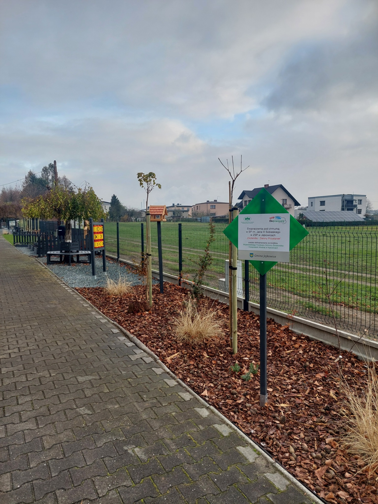 Nowo posadzone drzewka wzdłuż chodnika, zabezpieczone palikami, z rabatą wysypaną korą. Widoczny znak informacyjny o dofinansowaniu projektu.