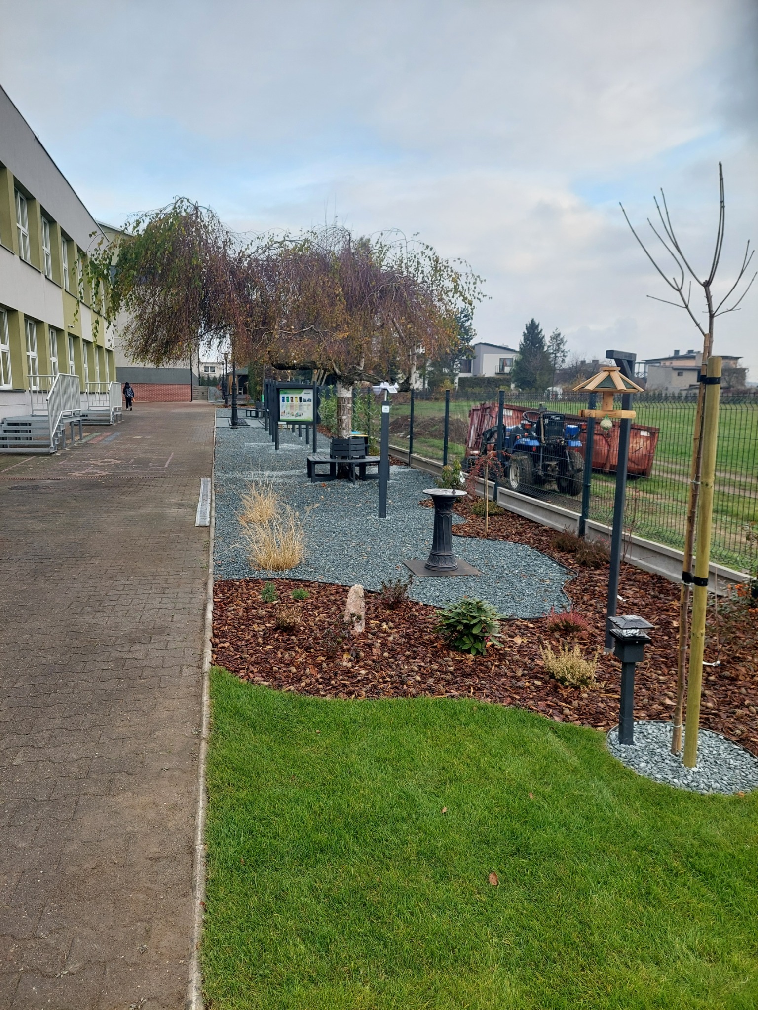 Uporządkowany teren zielony przy budynku szkolnym z elementami dekoracyjnymi: kora, kamień ozdobny, lampy solarne, karmnik dla ptaków, z tyłu widoczny traktor z przyczepą.