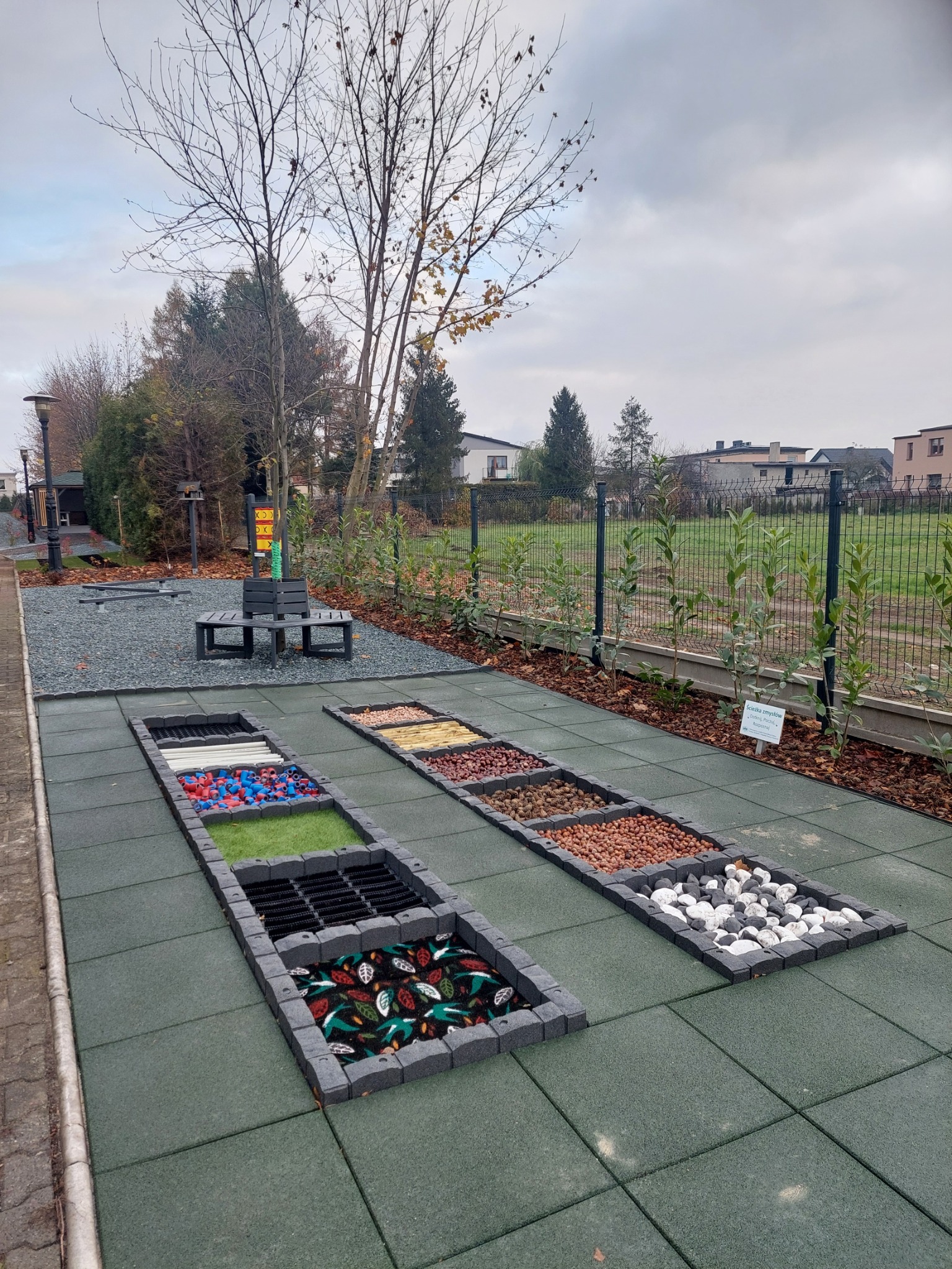 Ścieżka sensoryczna z różnymi powierzchniami: kamienie, żwir, kora, sztuczna trawa, drewno, tkanina z motywem liści, na gumowej nawierzchni placu zabaw.