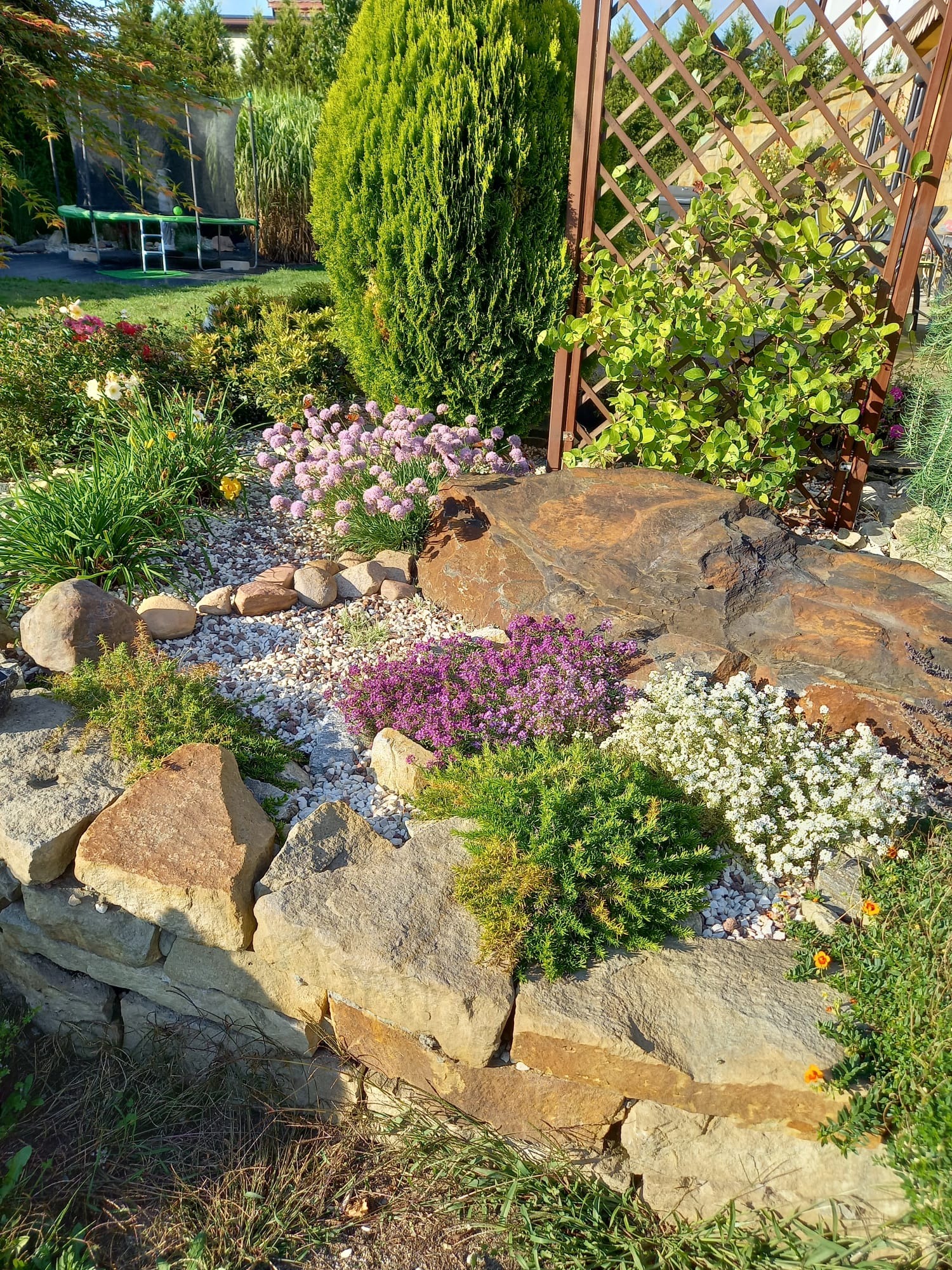 Ogród ozdobny z kamienną rabatą, różnokolorowymi kwiatami, krzewami i ozdobnym trejażem w tle.