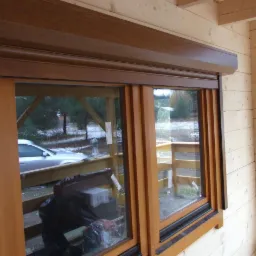 Brązowa roleta zewnętrzna zamontowana na drewnianym domu. Widoczne okno z drewnianą ramą i odbicie otoczenia w szybie. Zimowy krajobraz w tle.