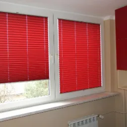 Dwa okna z zamontowanymi, czerwonymi plisami w pokoju z czerwonymi szafkami. Częściowo zasłonięte okno z widokiem na zewnątrz.