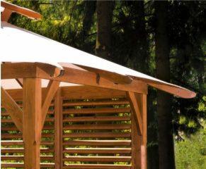 Drewniana altana ogrodowa z białym dachem, widoczna z bliska, z elementami ażurowych ścian i wspornikami dachu, na tle drzew.