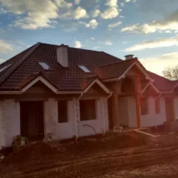 Dom w trakcie budowy z brązowym dachem, widoczne okna dachowe, rynny i komin. Budynek otoczony ziemią, w tle drzewa i niebo z chmurami o zachodzie słońca.