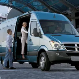 Niebieski bus Dodge Sprinter z otwartymi drzwiami przed wejściem do budynku, pasażerowie wsiadają, tropikalne rośliny w tle.