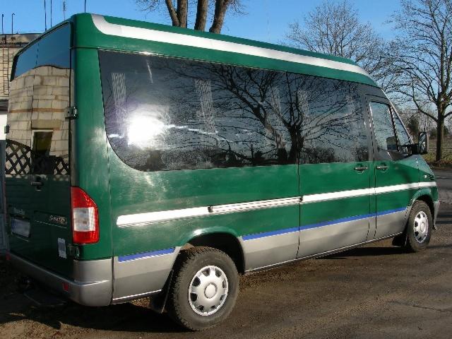 Zielony bus z białymi i szarymi akcentami, zaparkowany na żwirowym podłożu, z widocznym budynkiem z cegły w tle i odbiciem drzew na szybach.