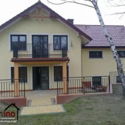 Żółty dom murowany z balkonem w Stadnikach, widok z ogrodu, czerwona dachówka, czarne balustrady, jasna kostka brukowa na podjeździe, logo firmy budowlanej w lewym dolnym rogu.