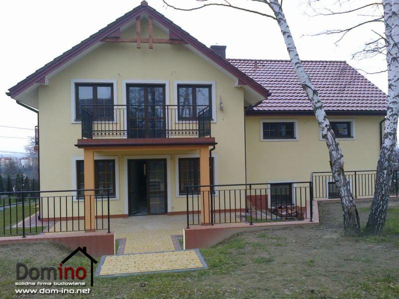 Żółty dom murowany z balkonem w Stadnikach, widok z ogrodu, czerwona dachówka, czarne balustrady, jasna kostka brukowa na podjeździe, logo firmy budowlanej w lewym dolnym rogu.