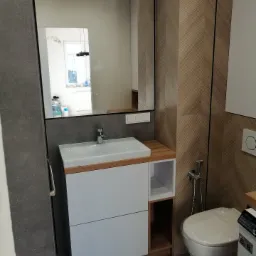 Nowoczesna łazienka z umywalką nablatową na szafce z otwartą p&oacute;łką, duże lustro w czarnej ramie i toaleta wisząca z bidetką, wykończona płytkami w geometryczny wz&oacute;r w odcieniach brązu.