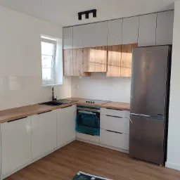 Nowoczesna kuchnia w bieli i drewnie: zabudowa z szarymi szafkami górnymi, drewnianymi frontami i blatem. Piekarnik zabezpieczony folią, stalowa lodówka.