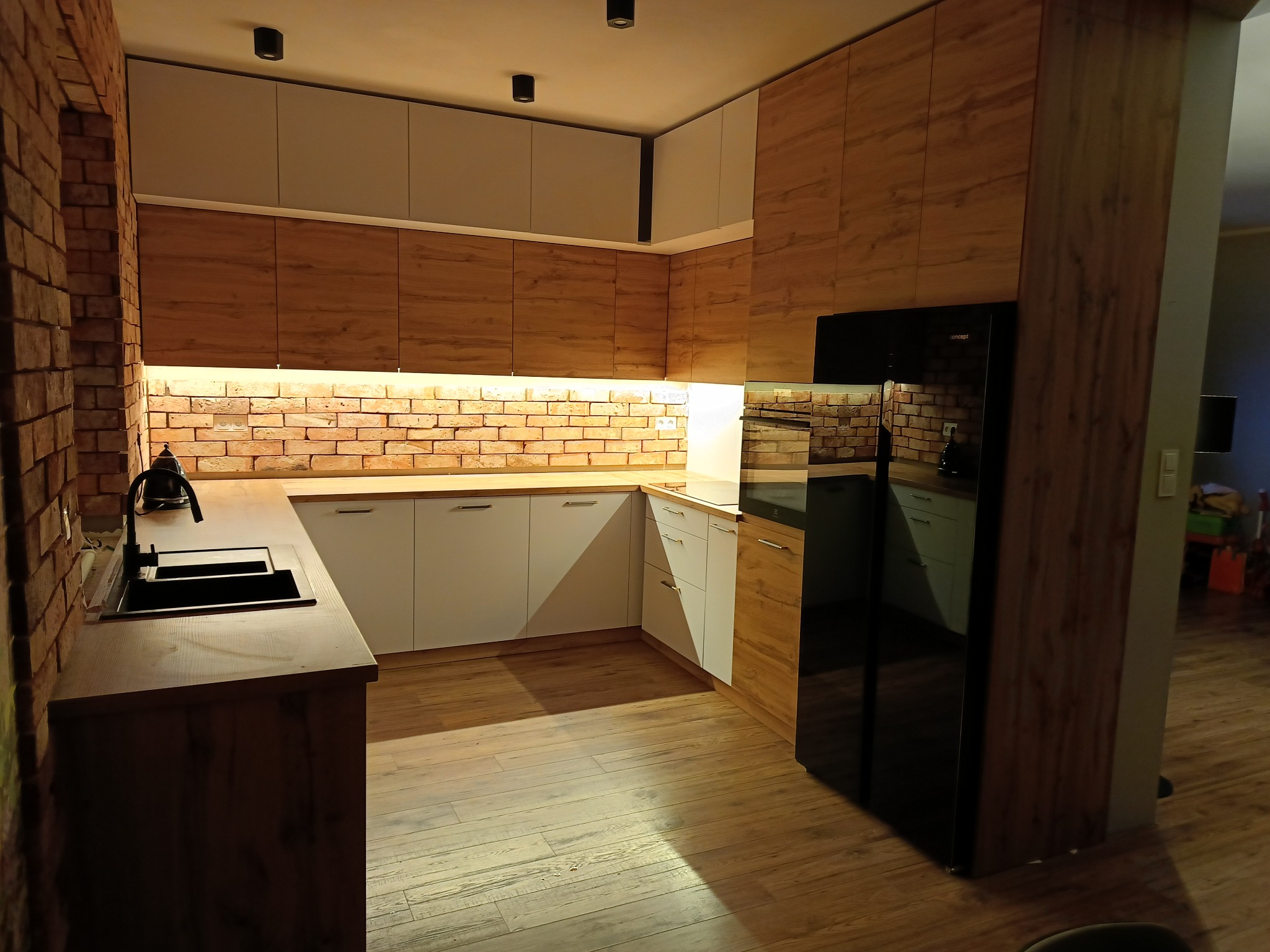 Nowoczesna kuchnia z ceglaną ścianą, drewnianymi i białymi szafkami oraz czarną lodówką. Ciepłe oświetlenie podkreśla fakturę drewna i cegły.