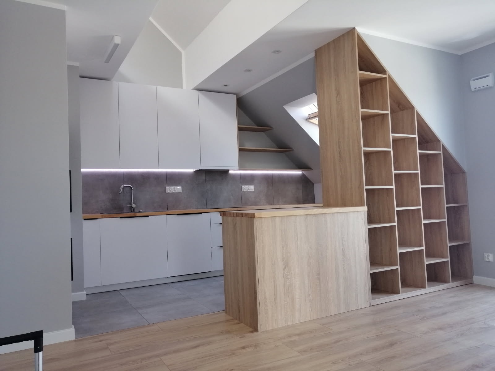 Biała kuchnia w skosach poddasza z drewnianą zabudową regałową, oświetleniem LED i szarą podłogą. Nowoczesna aranżacja wnętrza z elementami drewna i minimalizmu.