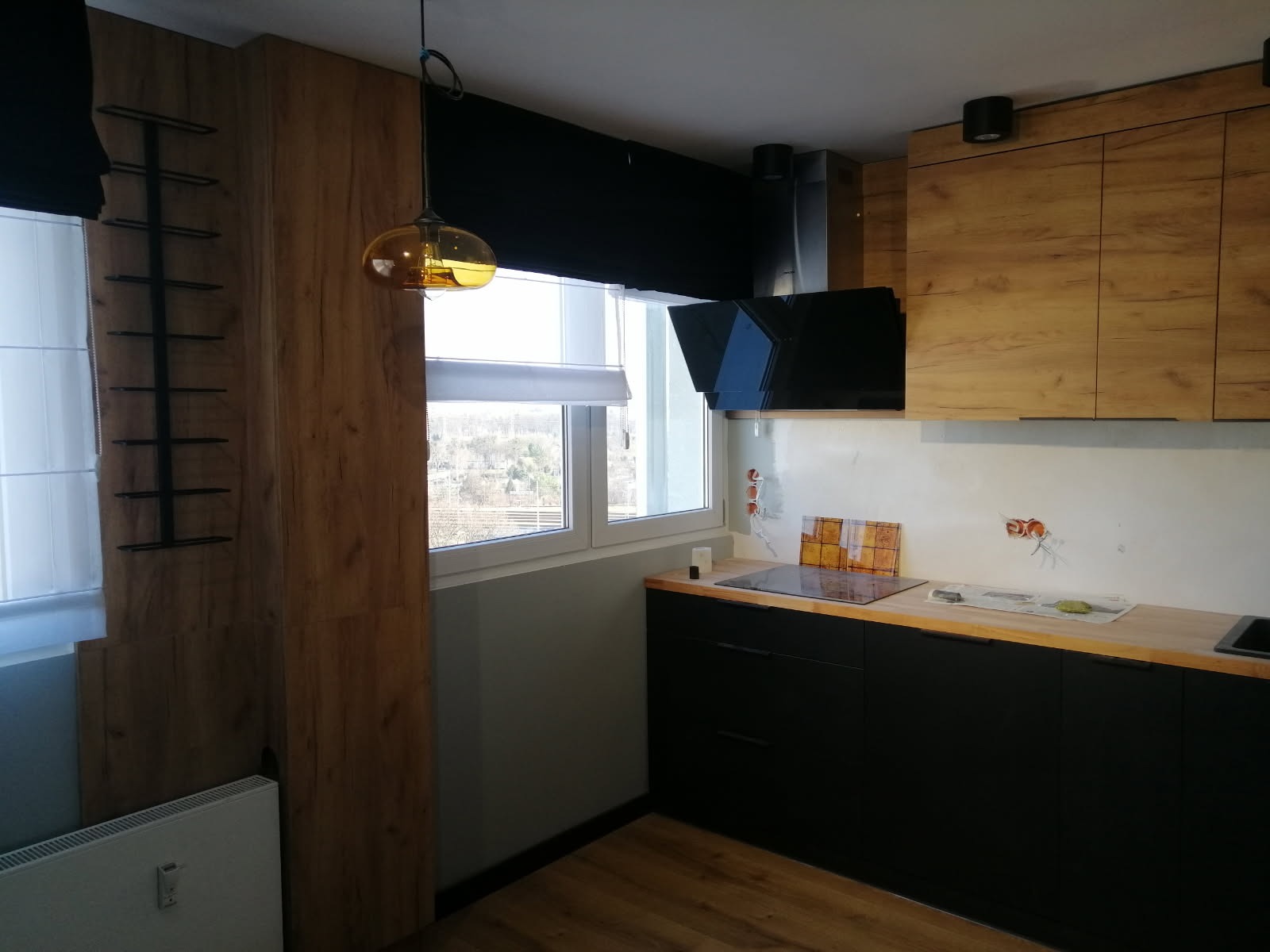 Nowoczesna kuchnia z drewnianymi szafkami, czarnymi frontami i blatem. Widoczny okap, okno z roletą i lampa wisząca. Ściana z widocznymi puszkami elektrycznymi.