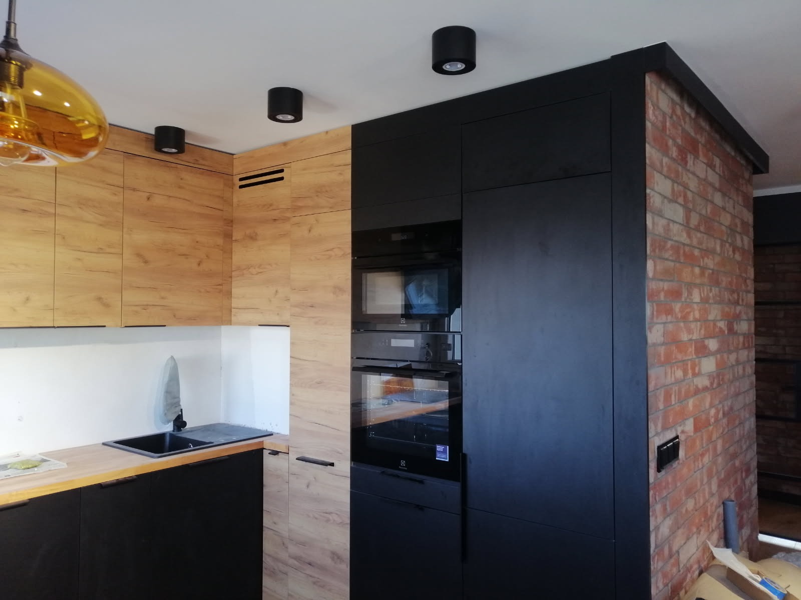 Nowoczesna kuchnia z zabudową w odcieniach drewna i czerni, oświetleniem punktowym i ceglaną ścianą w tle. Widoczny zlew i sprzęt AGD.