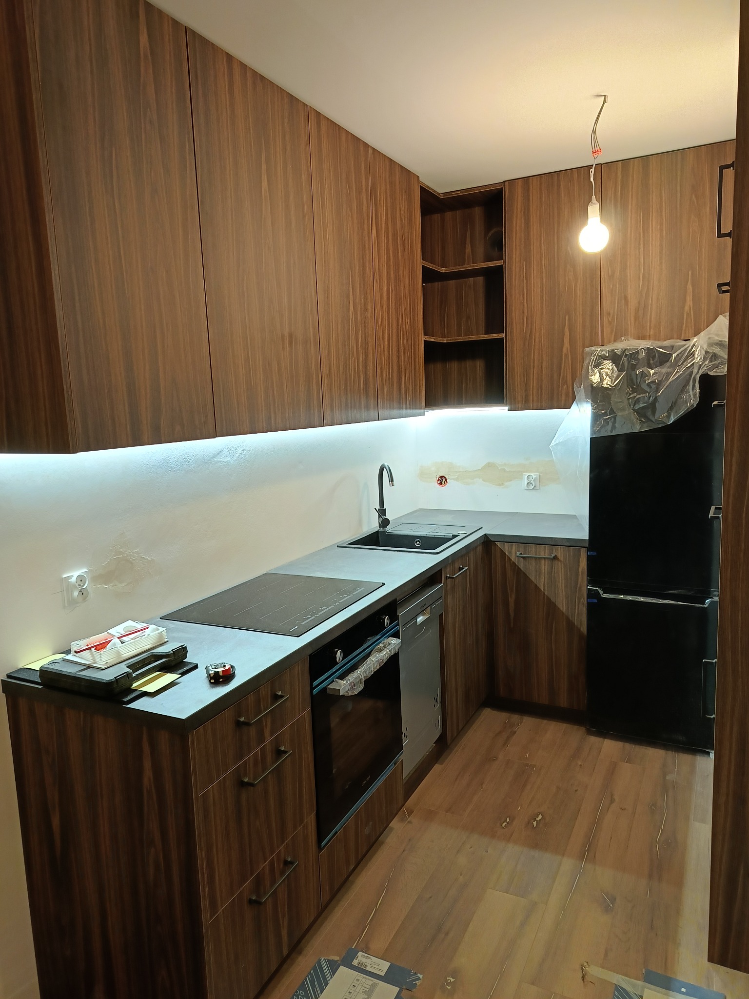 Nowoczesna kuchnia z drewnianymi szafkami, czarnym blatem i sprzętem AGD. Widoczne narzędzia na blacie, surowy fragment ściany z instalacją elektryczną.