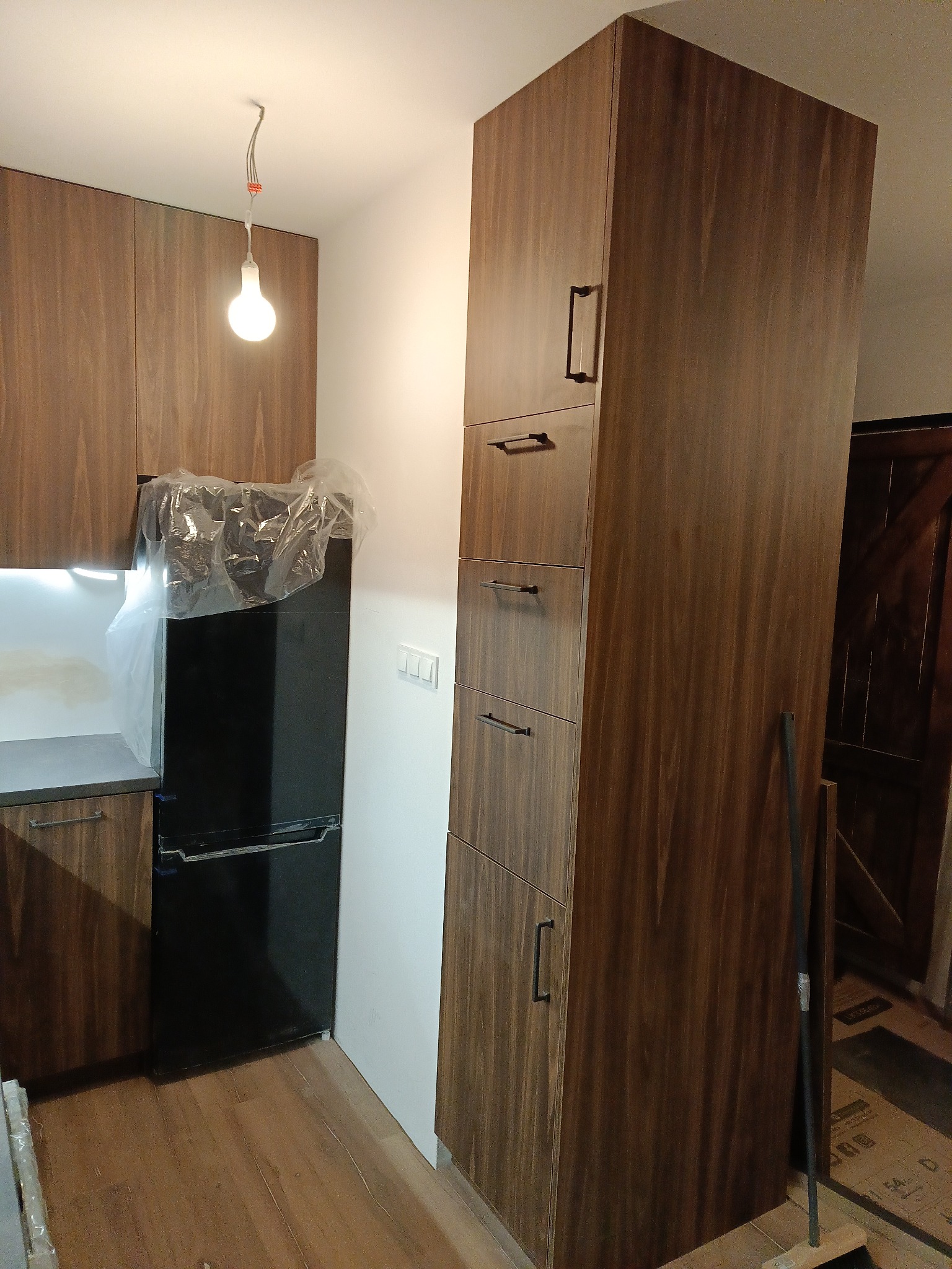 Nowoczesna kuchnia z drewnianymi frontami, czarną lodówką i jasnym oświetleniem. Wysoka zabudowa szafkowa z podłużnymi uchwytami. Podłoga drewniana.