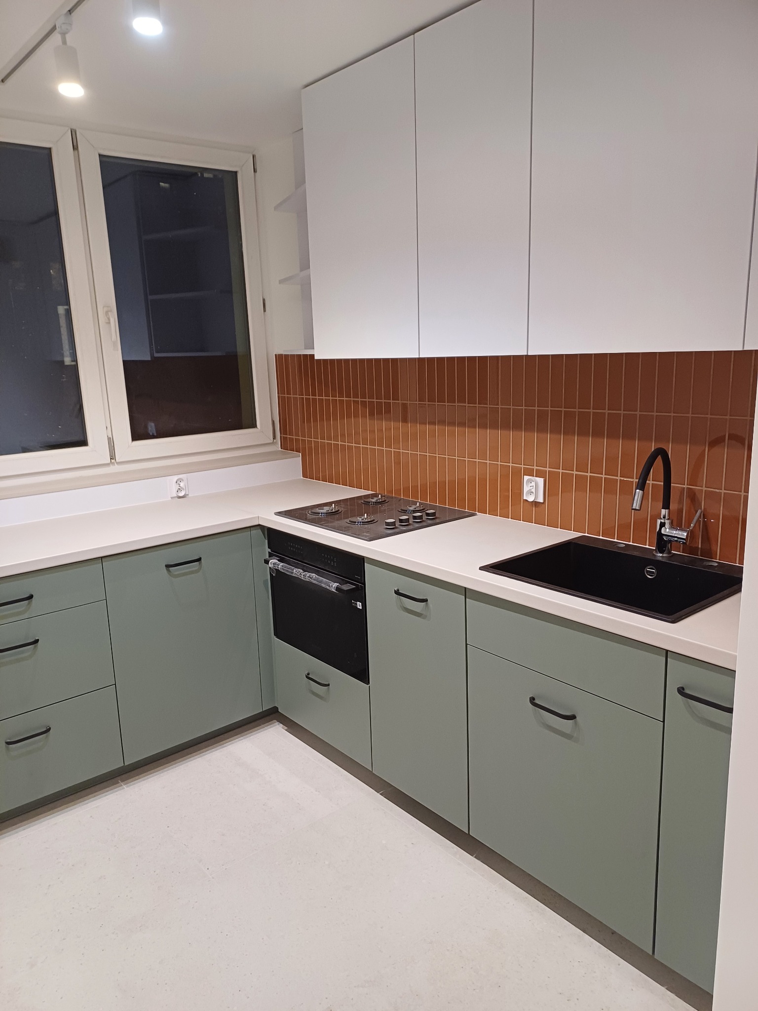 Nowoczesna kuchnia w odcieniach zieleni i bieli z brązowymi płytkami nad blatem. Minimalistyczne uchwyty, czarny zlew i płyta grzewcza. Oświetlenie sufitowe.