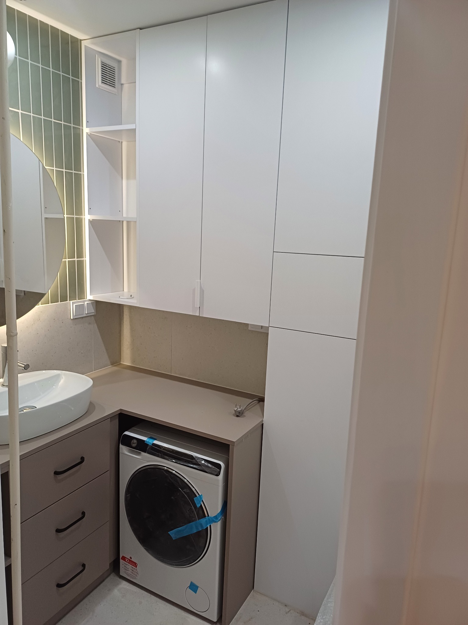 Nowoczesna łazienka z zabudową meblową: białe szafki wiszące, szara zabudowa pralki i umywalki z szufladami, okrągłe lustro z podświetleniem.