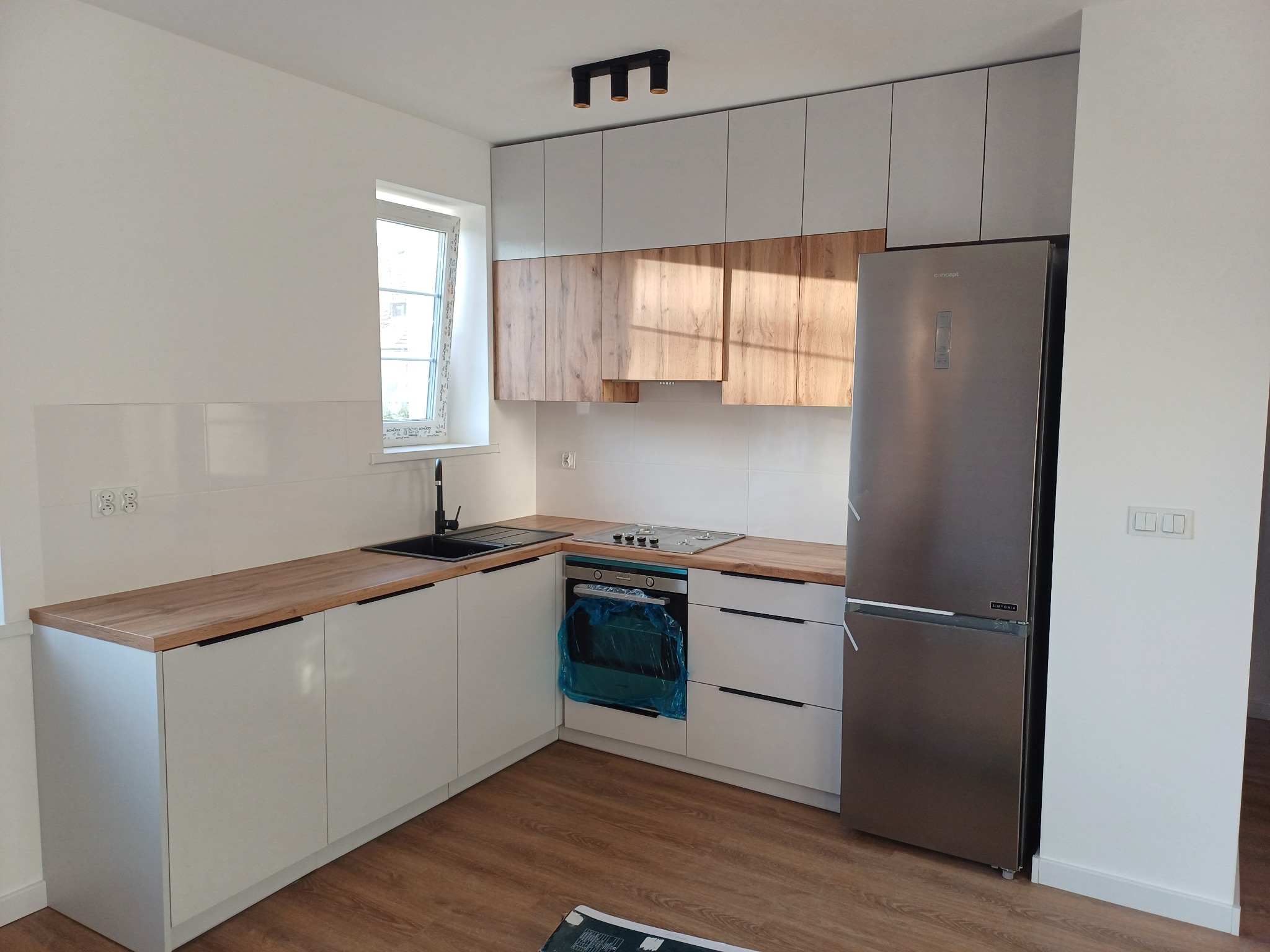 Nowoczesna kuchnia w bieli i drewnie: zabudowa z szarymi szafkami górnymi, drewnianymi frontami i blatem. Piekarnik zabezpieczony folią, stalowa lodówka.