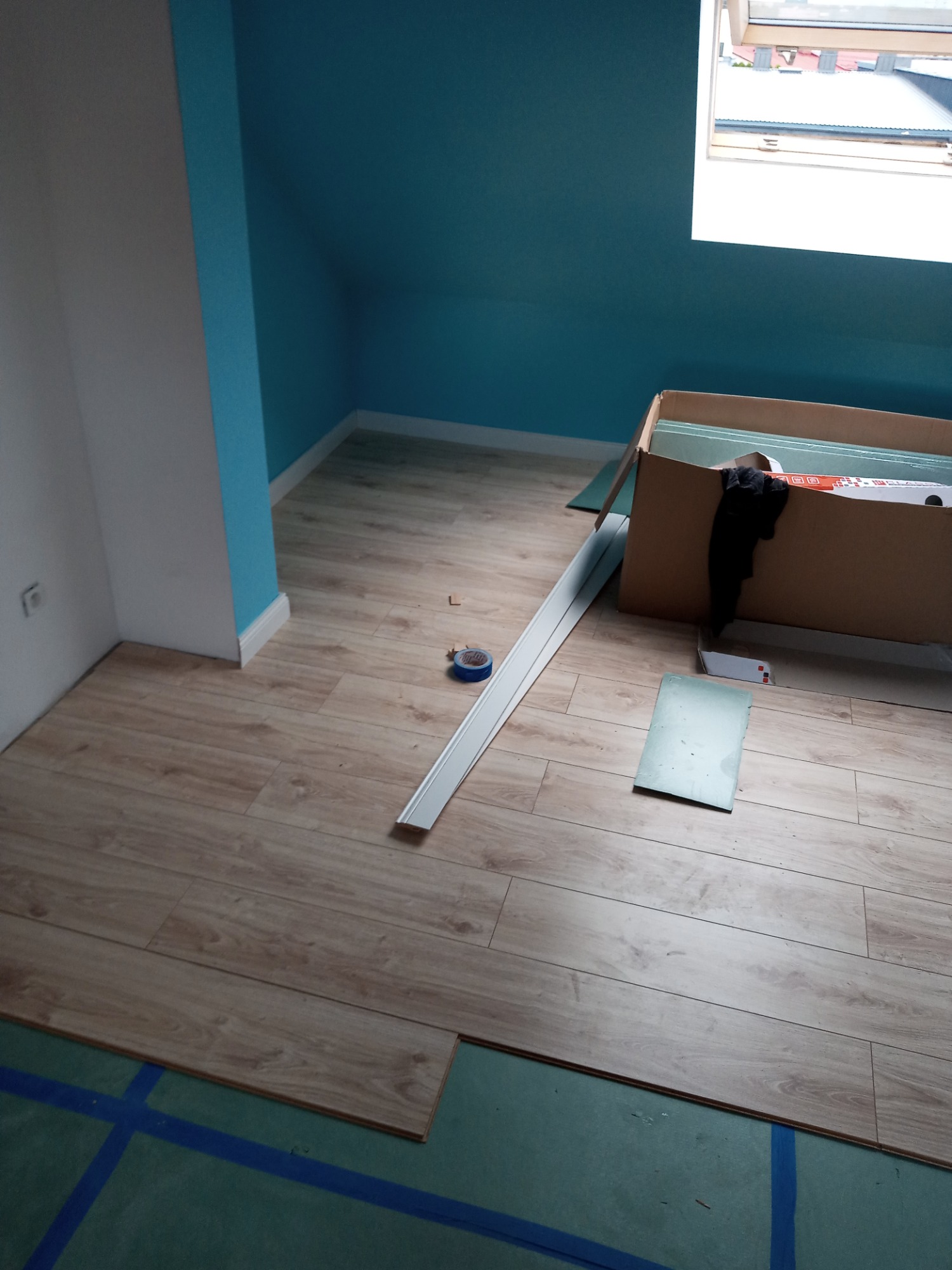 Układanie paneli podłogowych w pokoju na poddaszu, widoczne panele, listwa przypodłogowa, karton z panelami i niebieska taśma na podkładzie.