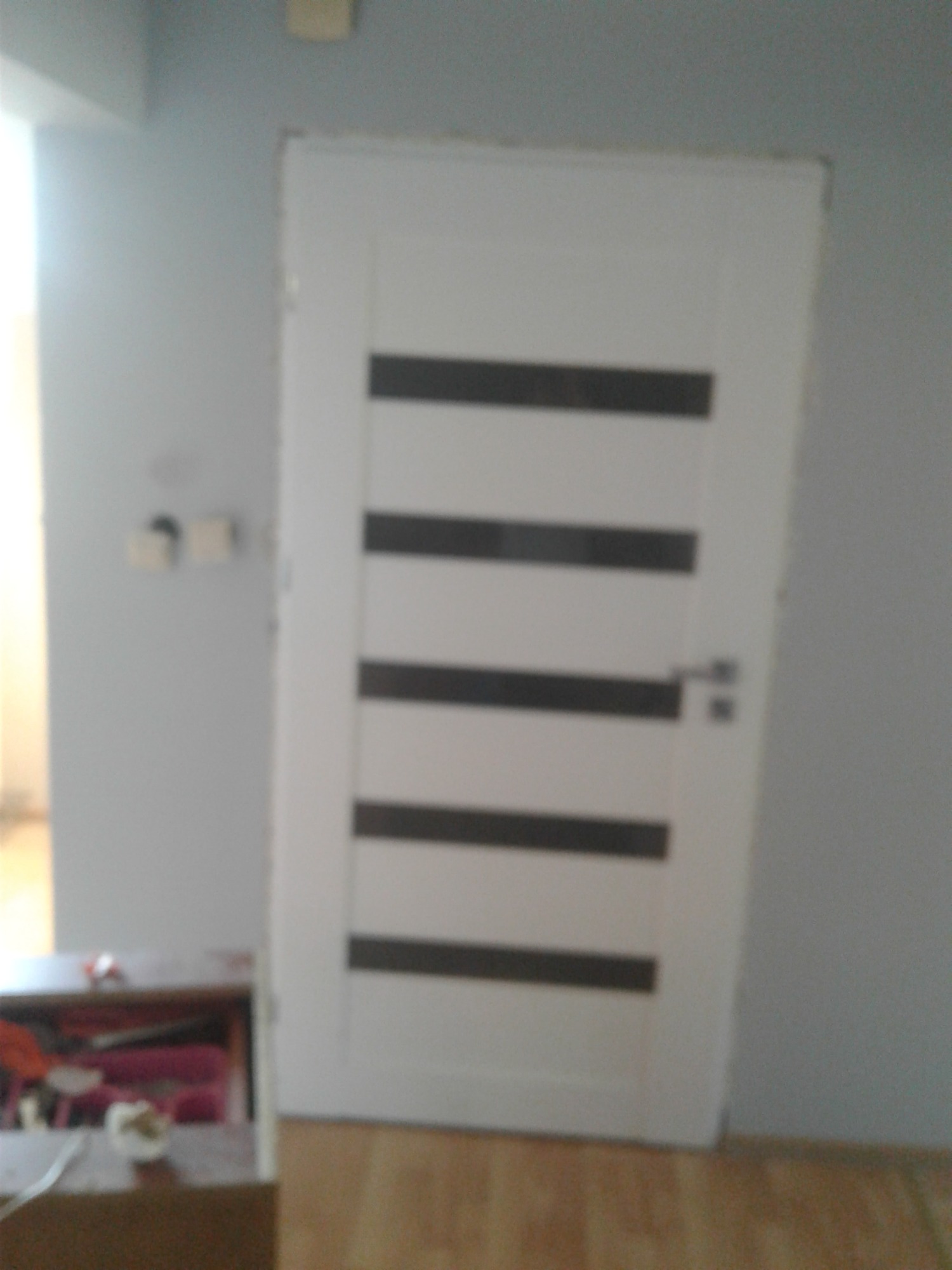 Białe drzwi wewnętrzne z poziomymi, ciemnymi przeszkleniami, zamontowane w szarej ścianie, widoczne fragmenty podłogi i stolika.