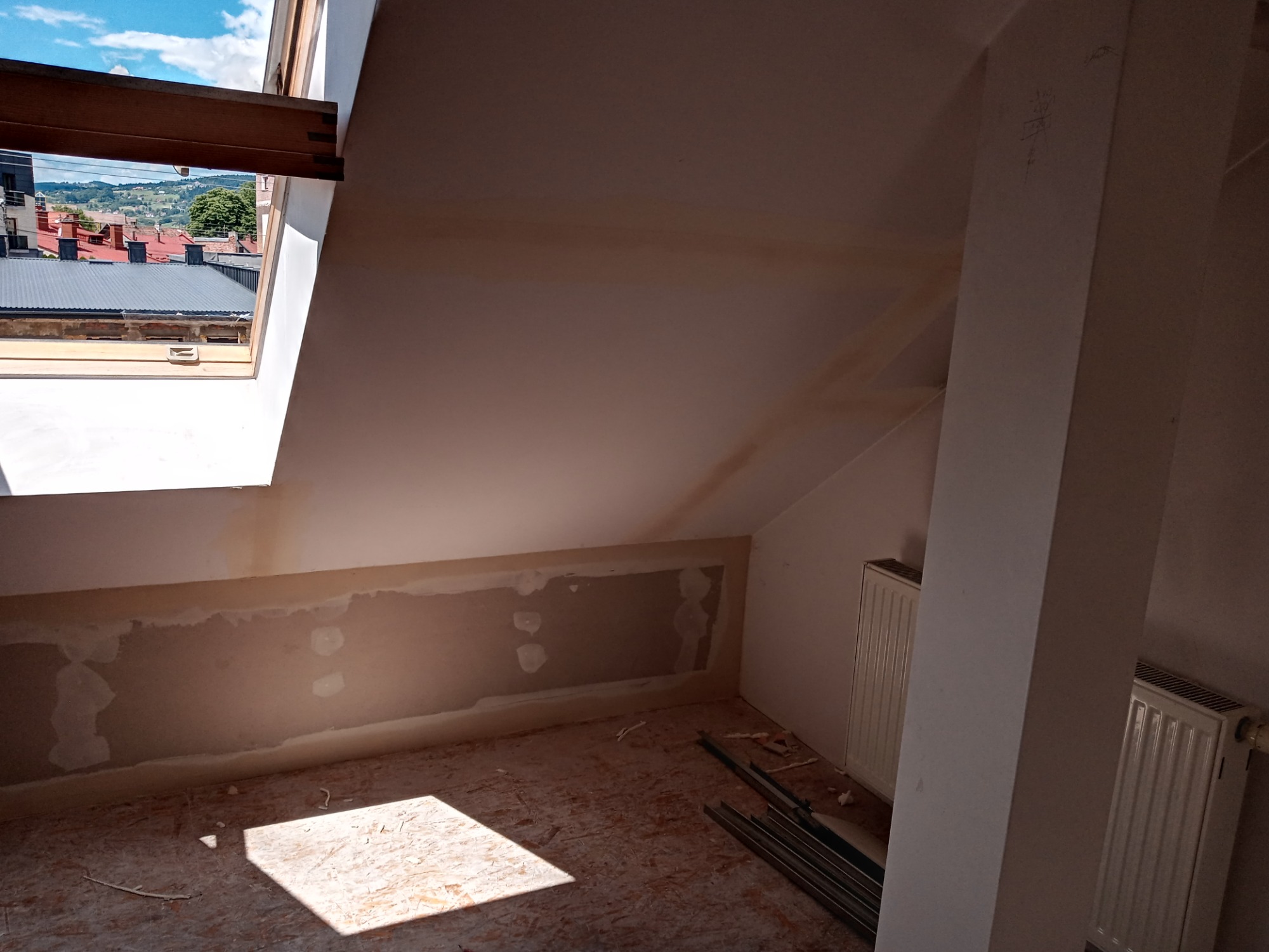 Poddasze w trakcie przygotowania do prac wykończeniowych: widoczne płyty kartonowo-gipsowe na ścianie, okno dachowe z widokiem na miasto, grzejniki i metalowe profile na podłodze.