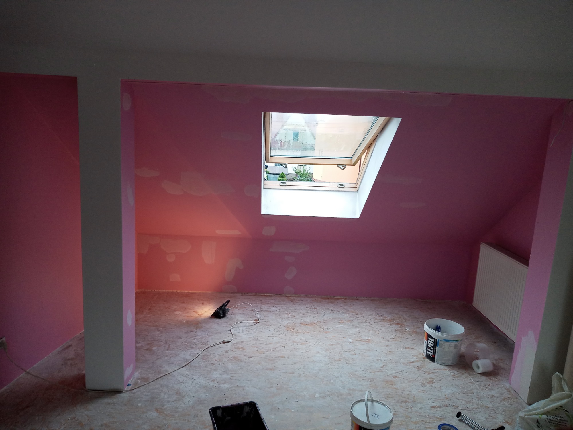 Poddasze w trakcie malowania na różowo, widoczne białe plamy po szpachlowaniu, otwarte okno dachowe, wiadro z farbą Alpol i narzędzia malarskie na podłodze.