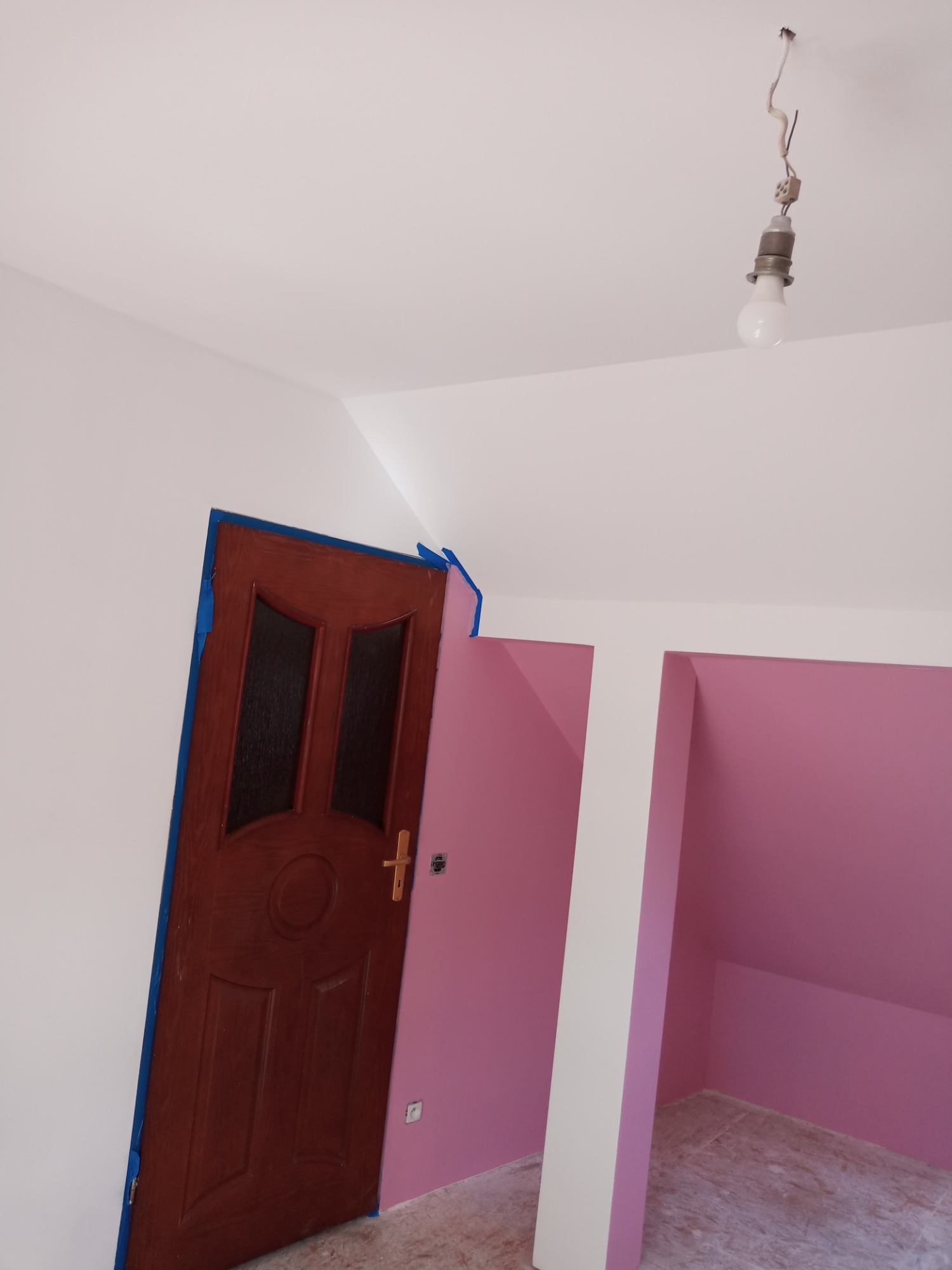 Pokój w trakcie odświeżania: ściany pomalowane na różowo, biały sufit z wiszącą żarówką, drewniane drzwi zabezpieczone niebieską taśmą malarską.