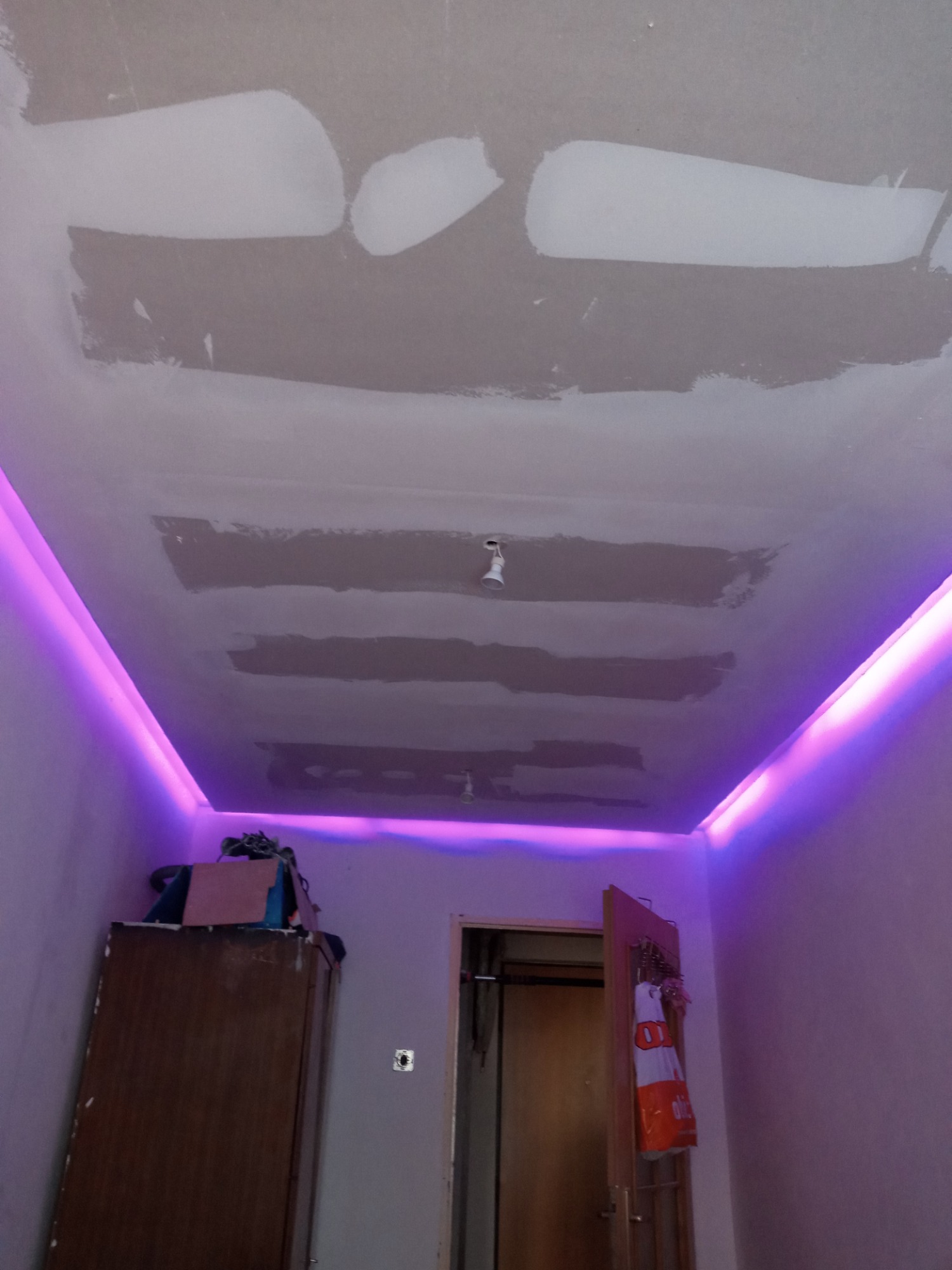 Niedokończony sufit podwieszany z widocznymi łączeniami płyt gipsowo-kartonowych, oświetlony fioletową listwą LED przy ścianach, widoczna szafa i drzwi w tle.