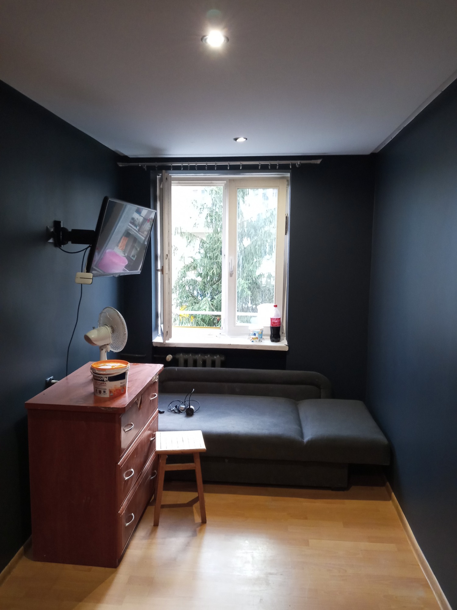 Pokój w Nowym Sączu po odświeżeniu ścian farbą w odcieniach grafitu, widoczna puszka z farbą na komodzie, mały taboret i rozłożona sofa.