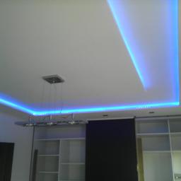 Darpol - Nowoczesny sufit podwieszany z oświetleniem LED w kolorze niebieskim, widoczny fragment zabudowy meblowej z półkami i wisząca lampa. Aranżacja wnętrza.