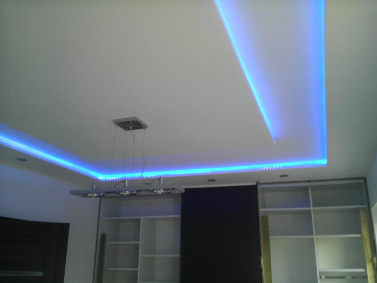 Nowoczesny sufit podwieszany z oświetleniem LED w kolorze niebieskim, widoczny fragment zabudowy meblowej z półkami i wisząca lampa. Aranżacja wnętrza.