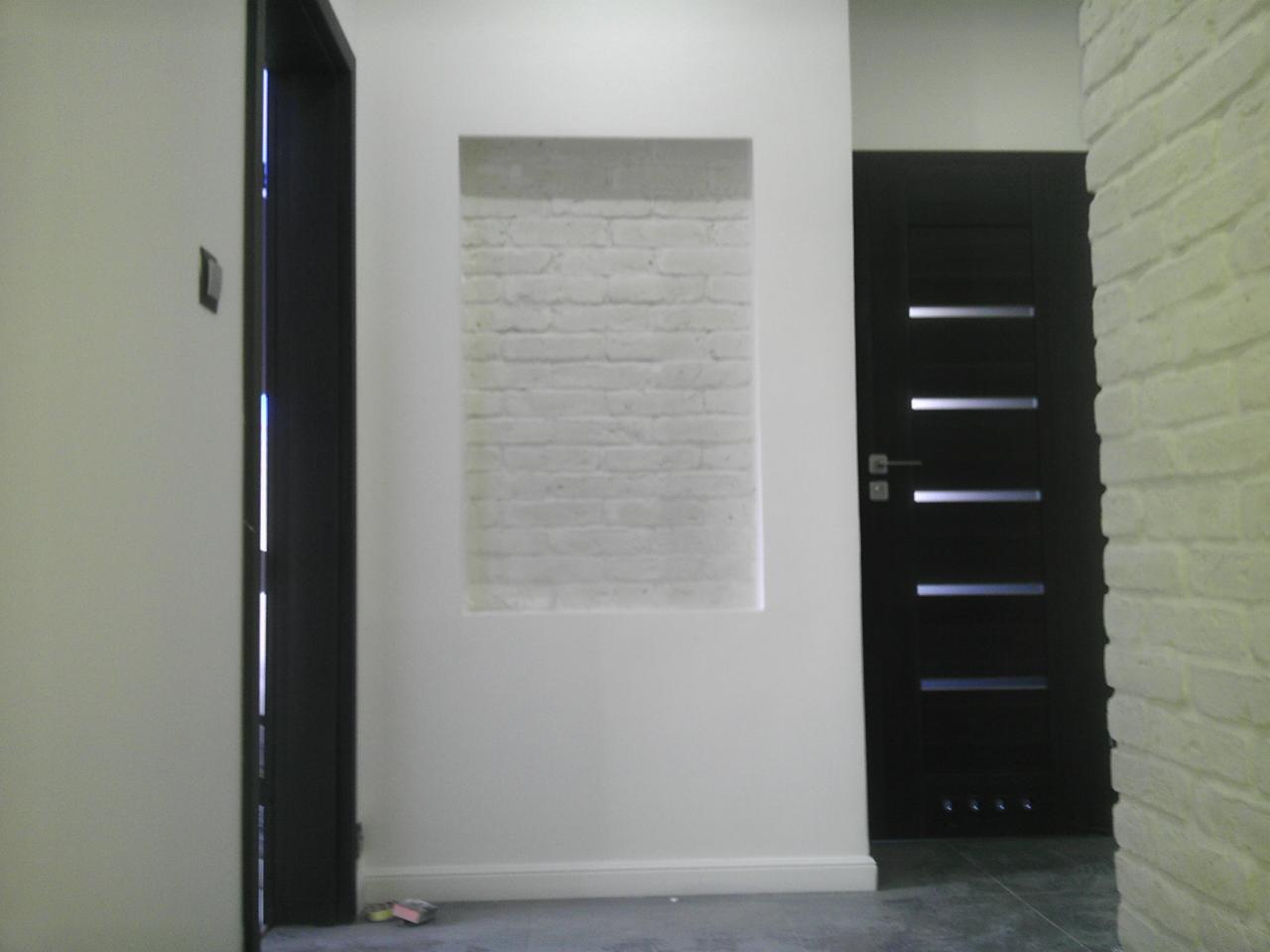 Wnęka z białej cegły w ścianie, obok czarne drzwi z przeszkleniami i drugie drzwi po lewej stronie. Wykończenie wnętrza w nowoczesnym stylu.